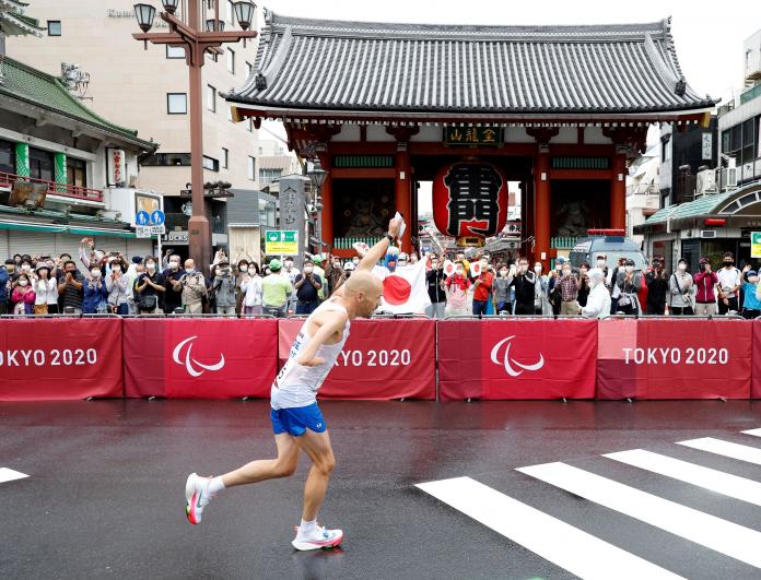 A male marathon runner lifts his hand up in a salute as he runs past spectators on a wet cement road.